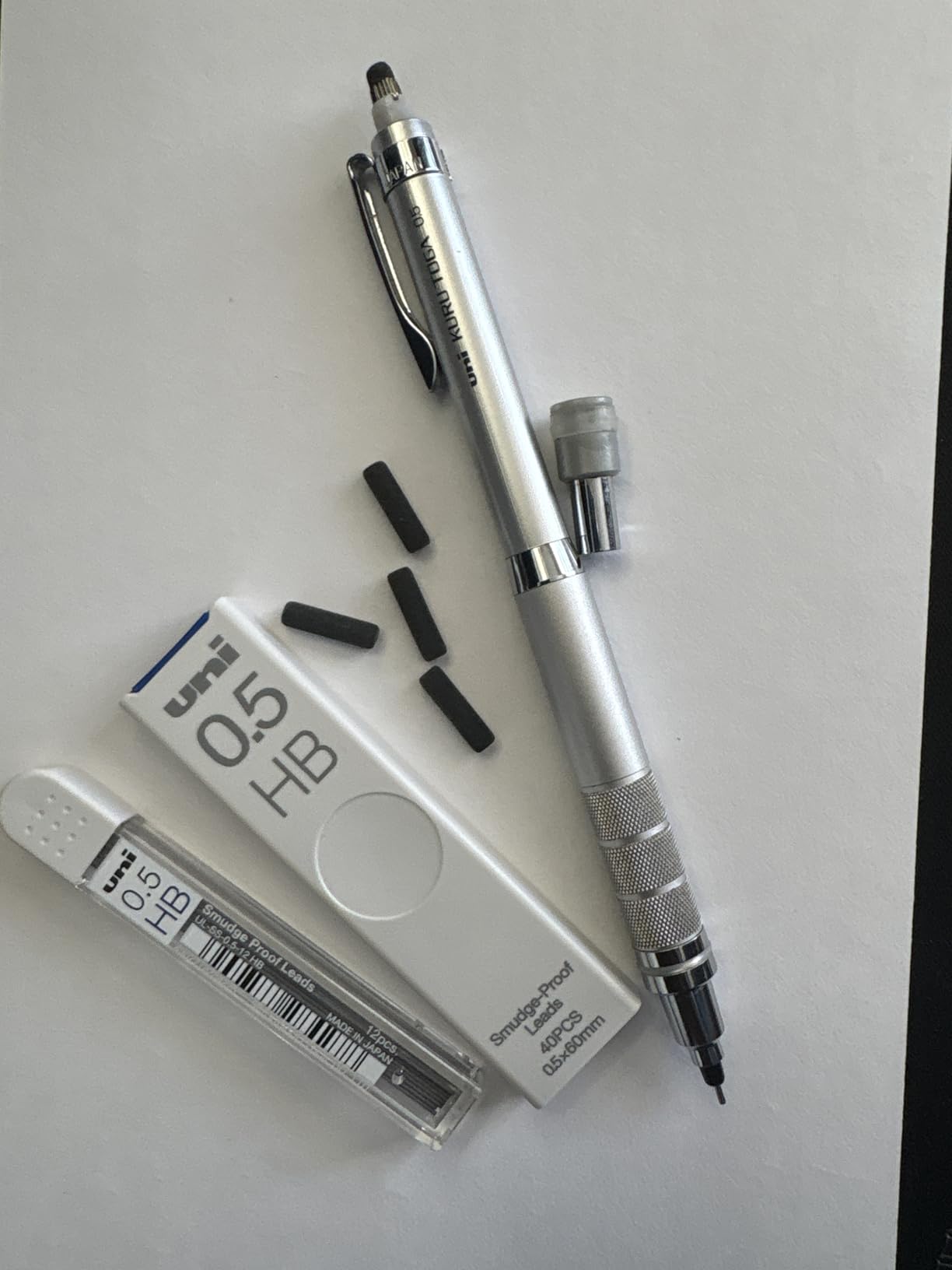 Silver Kuru Toga Roulette on white paper beside two tubes of uni 0.5 HB lead and four spare black erasers tipped out of the cap