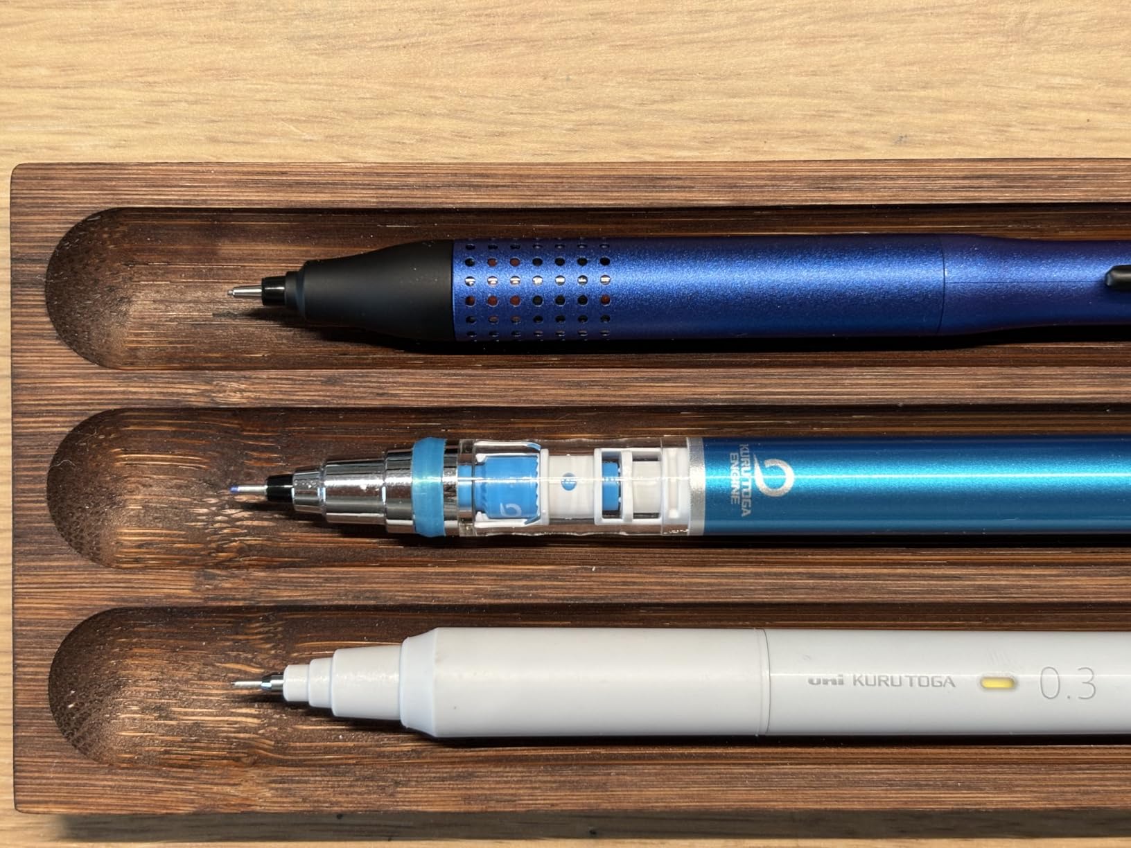 Three Kuru Toga pencils lined up in a wooden tray: a navy Advance Upgrade, a blue Advance with its clear engine housing exposed, and a white 0.3 mm model