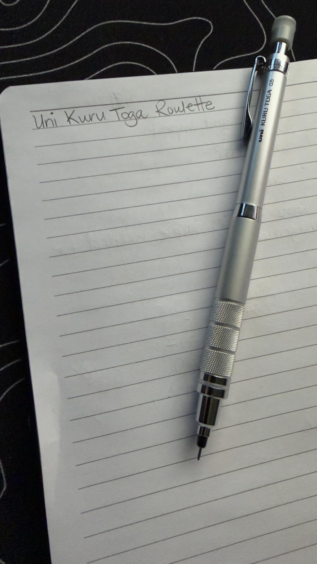 Silver Kuru Toga Roulette lying on a lined notepad with Uni Kuru Toga Roulette written across the top line in pencil