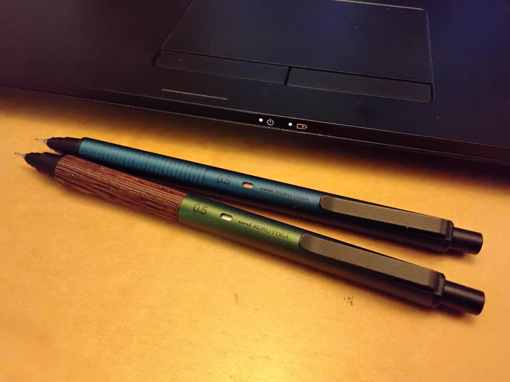 A teal Kuru Toga Metal and a Forest Green Kuru Toga Wood side by side on an orange desk in front of a laptop, both marked 0.5