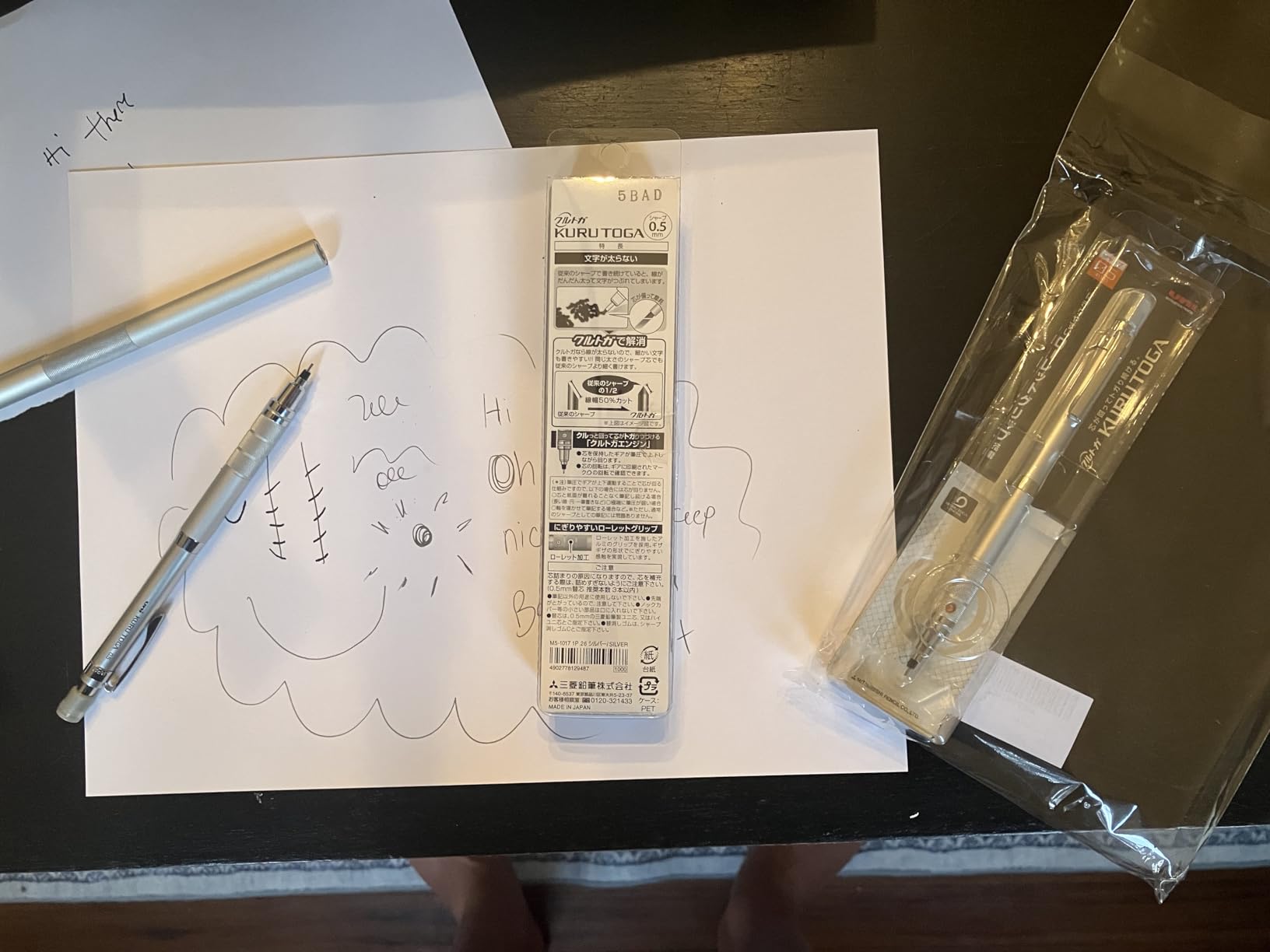 Silver Kuru Toga Roulette resting on a sheet of pencil doodles beside its Japanese packaging marked M5-1017 1P.26 and a second sealed pencil